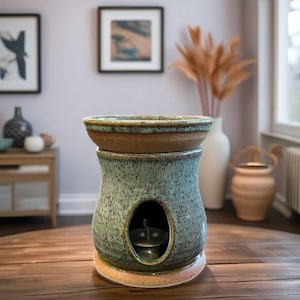 May include: A ceramic essential oil diffuser with a speckled teal glaze. The diffuser has a round top, a central opening, and a base with a brown rim. The design includes a small opening for a tea light. Home decor.