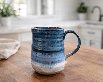 May include: A handmade ceramic mug with a blue and white glaze. The mug has a rounded body, a sturdy handle, and a textured surface. The mug is sitting on a wooden surface. The mug is suitable for coffee, tea, or other beverages.