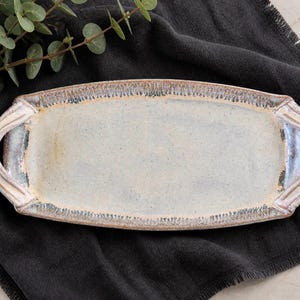 May include: A rectangular ceramic serving platter with handles. The platter has a light blue and cream colored surface with a textured rim. The handles are a cream color. The platter is on a dark gray cloth.