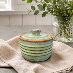May include: A round ceramic lidded jar featuring a light green and turquoise glaze with horizontal ribbed textures. The rim of the jar and the lid edge display a raw, dark brown clay finish, and the lid has a small, centered knob handle.