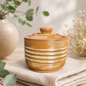 May include: A cylindrical ceramic jar with a matching lid, featuring a glossy mustard-yellow glaze and horizontal ribbed textures along the body. The vessel has a smooth, rounded knob on top of the lid and a consistent, warm-toned finish.
