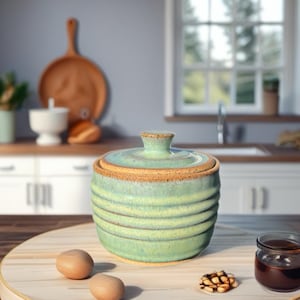 May include: A handcrafted, green ceramic jar with a lid, featuring horizontal ridges and a natural, earthy rim. The jar sits on a wooden tray with two eggs, a small jar of preserves, and a walnut. The background includes a kitchen setting.