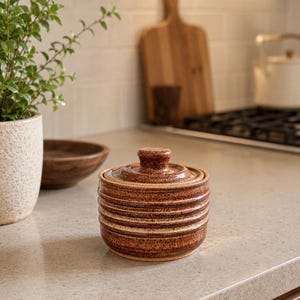 May include: A small, round, ceramic sugar bowl with a lid. The bowl is brown with horizontal bands of lighter brown and a small knob on the lid. The bowl sits on a light-colored countertop in a kitchen setting.