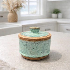 May include: A ceramic jar with a lid, featuring a speckled turquoise glaze and a natural brown rim. The jar is cylindrical with a small knob on top of the lid. The design is rustic and handmade, suitable for kitchen storage.
