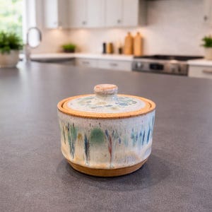 May include: A ceramic jar with a lid, featuring a speckled, light-colored glaze with blue, green, and red drips. The jar sits on a dark countertop in a kitchen setting. The lid has a small, round knob.