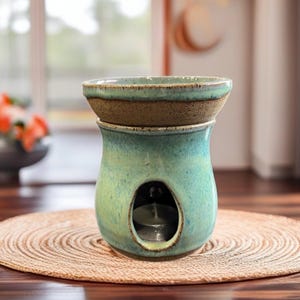 May include: A teal ceramic essential oil diffuser with a brown rim and a small opening for a tea light. The diffuser sits on a round, woven placemat. The design features a rounded base and a separate top dish.