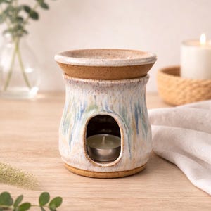 May include: A ceramic essential oil diffuser with a candle inside. The diffuser is white with blue and green accents and a brown rim. The candle sits in a metal container. The diffuser is on a wooden surface.