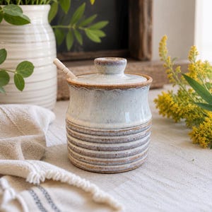 May include: A ceramic honey pot with a light blue and white speckled glaze, featuring a matching lid and a small wooden dipper. The base of the pot has horizontal ribbed textures, while the rim displays a subtle wavy pattern in brown tones.