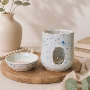 May include: A ceramic wax warmer with a speckled light blue and white glaze featuring a large oval cutout for a tea light candle, accompanied by a matching small speckled bowl containing pink wax melts on a round wooden tray.