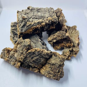 May include: A pile of rough, textured cork bark pieces. The bark has a natural, earthy color palette with shades of brown, gray, and tan. The pieces vary in size and shape, showcasing the organic texture of the bark.