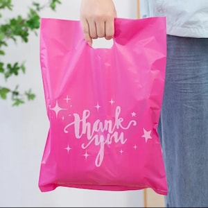 May include: A bright pink plastic shopping bag with a die-cut handle. The bag features the words "thank you" in white script, along with star and sparkle graphics. The bag is held by a person.