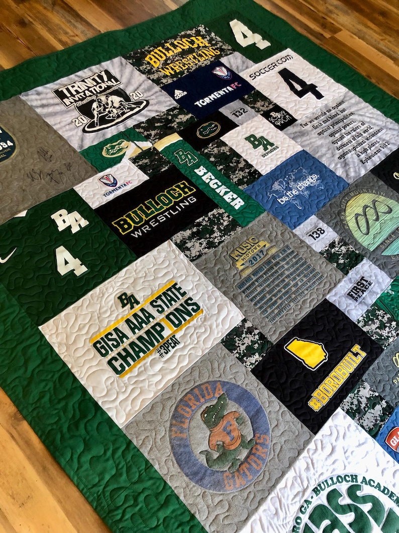 以下が含まれることがあります： 緑、白、グレーのパッチワークキルトは、Tシャツで作られています。キルトには、「Bulloch Wrestling」、「GISA AAA State Champ Ons」、「Tormenta FC」、「Be the Change」など、さまざまなロゴとテキストが描かれています。