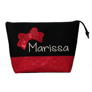 Puede incluir: Bolsa negra con cremallera y fondo de purpurina roja. La bolsa tiene un lazo rojo de lentejuelas y el nombre "Marissa" bordado en blanco.