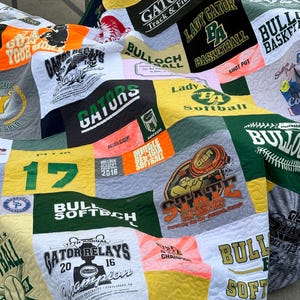 以下が含まれることがあります： 緑、白、黄色のパッチワークキルトは、古いTシャツで作られています。キルトには、「ゲーター」、「バロックアカデミー」、「レディーゲーター」、「ソフトボール」、「ブル」、「ステート」など、さまざまなスポーツチームのロゴと名前が描かれています。