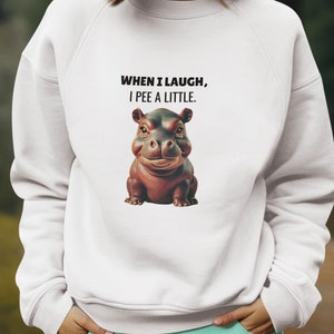 Puede incluir: Sudadera blanca con un dicho divertido y un hipopótamo de dibujos animados. El texto dice "When I laugh, I pee a little."