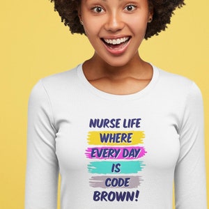 Peut inclure: Un t-shirt blanc à manches longues avec le texte "Nurse Life Where Every Day Is Code Brown!" imprimé sur le devant. Le texte est en lettres colorées de style coup de pinceau.