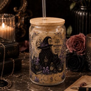 May include: A clear glass tumbler with a bamboo lid and a glass straw, featuring a printed graphic of a witch in a hat, a black cat, crystals, and the text: She works in mysteries and makes magic, surrounded by floral and celestial motifs.