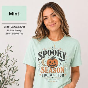 Op de afbeelding: Een mintgroen t-shirt met korte mouwen en een grafische print met de tekst "SPOOKY SEASON SOCIAL CLUB" en "GOOD VIBES - CREEPY TIMES EST. 2026". Het ontwerp bevat een centrale pompoen, een maansikkel, vleermuizen en sterren.
