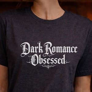 以下が含まれることがあります： 白い装飾的なフォントで「Dark Romance Obsessed」というテキストがプリントされた濃い灰色のTシャツ。クルーネックのTシャツで、柔らかい素材で作られているようです。全体的な雰囲気は、文学的またはゴシック的なテーマを示唆しています。
