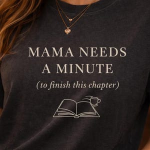 Può includere: T-shirt grigio scuro con la scritta bianca "MAMA NEEDS A MINUTE (to finish this chapter)". La maglietta presenta un disegno a tratto di un libro aperto con una tazza di caffè. È un capo casual.