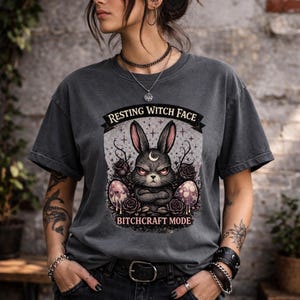 May include: Dark gray t-shirt with a graphic of an angry bunny with a crescent moon on its forehead, surrounded by roses and eggs. Text reads "RESTING WITCH FACE" above and "BITCHCRAFT MODE" below the graphic.