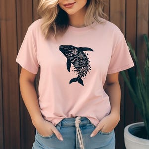 Könnte beinhalten: Hellrosa T-Shirt mit einem schwarzen Wal-Design, gefüllt mit floralen und Wellenmustern. Das T-Shirt hat kurze Ärmel und einen Rundhalsausschnitt. Das Wal-Design befindet sich mittig auf der Vorderseite.