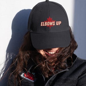 Puede incluir: Una gorra de béisbol negra con una hoja de arce roja y el texto "Elbows Up" bordado en la parte delantera.