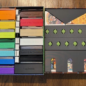 Könnte beinhalten: Ein Brettspiel-Organizer-Einsatz mit zwei rechteckigen Boxen, gefüllt mit beschrifteten Kartenhaltern aus Kunststoff in Orange, Gelb, Grün, Blau, Lila, Rot, Beige, Weiß und Schwarz, sowie einem dunkelgrauen Tablett mit zwölf kleinen grünen runden Spielsteinen.