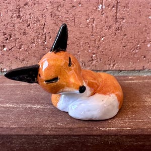 May include: A small ceramic fox figurine in a curled, sleeping position. The fox features a glossy orange coat, black-tipped ears, closed eyes, and a white chest and tail tip. The sculpture has a smooth, reflective finish and a small black nose.