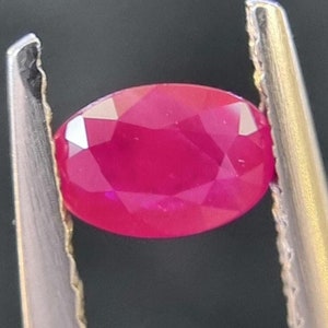 May include: A vibrant red oval-shaped gemstone held in a pair of silver tweezers.