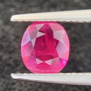 May include: A vibrant pink oval-shaped gemstone with a faceted cut, held by tweezers against a dark grey background.