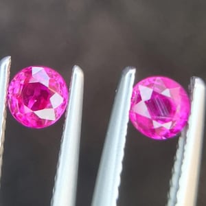 以下が含まれることがあります： 2つの丸いピンクの宝石がピンセットで保持されています。宝石はファセットカットされており、明るく光沢のある仕上がりです。