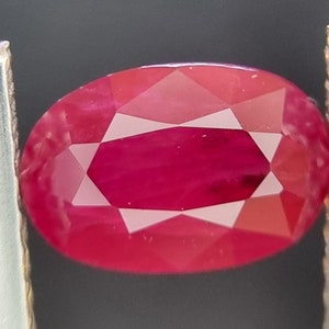 Puede incluir: Una gema ovalada de color rojo vibrante con un corte facetado, sostenida por unas pinzas.