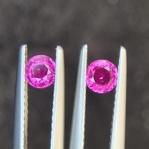 Könnte beinhalten: Zwei runde, rosafarbene Edelsteine ​​werden in einer Pinzette gehalten. Die Edelsteine ​​sind facettiert und haben eine leuchtende, lebendige Farbe.