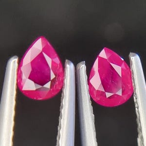 以下が含まれることがあります： 深い赤色のペアシェイプのルビーの宝石が2つ、ピンセットで保持されています。宝石はファセットカットされており、非常に高い輝きを持っています。