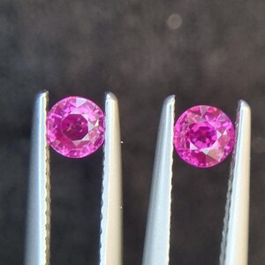 以下が含まれることがあります： 2つの丸いピンクの宝石がピンセットで保持されています。宝石はファセットカットされており、明るく鮮やかな色をしています。