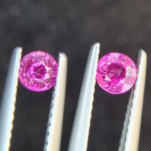 以下が含まれることがあります： 2つの丸い、鮮やかなピンク色の宝石が、銀色のピンセットで挟まれています。宝石は光を反射するファセット加工された表面を持っています。暗い背景は石の明るい色を強調しています。これらの宝石はおそらくジュエリー作りに使用されます。