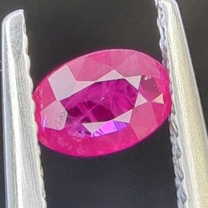 May include: A vibrant pink oval-shaped gemstone held in a pair of silver tweezers.