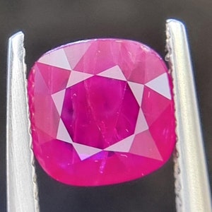 May include: A vibrant pink ruby gemstone with a faceted cut, held in a pair of tweezers.