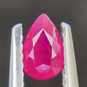 Pode incluir: Uma gema rosa vibrante em forma de pêra, segurada por uma pinça de prata.