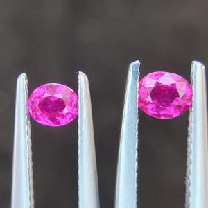 May include: Two oval-shaped pink gemstones are held in tweezers. The gemstones are a vibrant pink colour and have a smooth, polished surface.