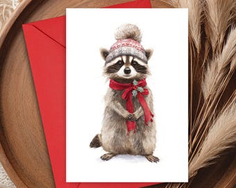 May include: A watercolour illustration of a raccoon wearing a red scarf and a grey and red knitted hat with a pom-pom. The raccoon is on a white card, with a red envelope, resting on a wooden tray. Decorative pampas grass is in the background.