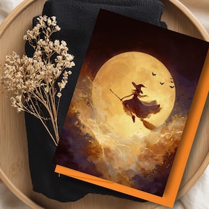 May include: A Halloween-themed greeting card featuring a witch flying on a broom in front of a full moon. The card has an orange border and is set on a wooden tray with dried flowers and a black cloth.