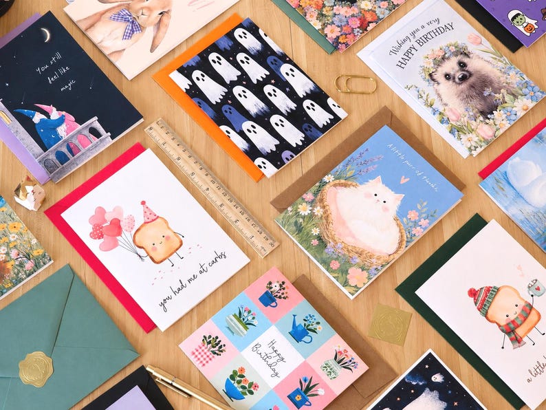 Op de afbeelding: Een verzameling rechthoekige wenskaarten met diverse illustraties waaronder spoken, een egel, een kat en toast-figuren met tekst zoals 'You still feel like magic', 'HAPPY BIRTHDAY' en 'you had me at carbs', naast een liniaal van 30 cm.