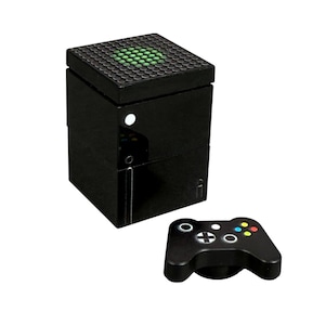 Peut inclure: Une console de jeu noire en forme de cube avec une manette assortie. La console présente un dessus texturé avec des lumières vertes et un bouton d'alimentation blanc. La manette a des boutons colorés et un pavé directionnel.