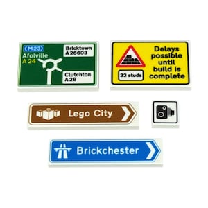 Op de afbeelding: Een verzameling Lego City-verkeersborden, waaronder een groen bord met een rotonde en de tekst "Bricktown A 26603", een geel bord dat waarschuwt voor vertragingen en een bruin bord met de tekst "Lego City".