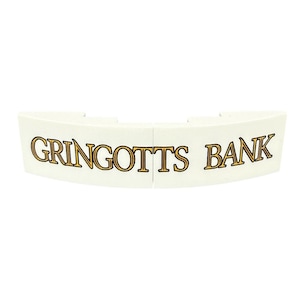 May include: Two curved white plastic building block pieces placed side by side. The pieces feature the text GRINGOTTS BANK printed in a stylized gold font with a thin black outline, spanning across the front surface of both connected segments.