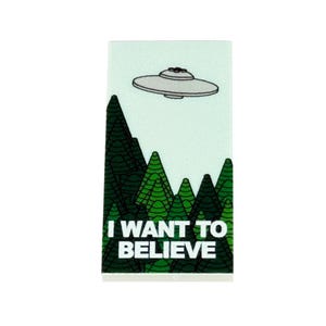 May include: A rectangular sticker with a light blue background features a gray UFO above green stylized trees. The text "I WANT TO BELIEVE" is printed in white at the bottom of the sticker.