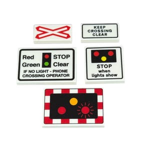 Op de afbeelding: Een verzameling witte, rechthoekige borden met zwarte randen, met verschillende symbolen en tekst gerelateerd aan het spoorwegverkeer. De borden bevatten teksten zoals "KEEP CROSSING CLEAR" en instructies met gekleurde lichten die stoppen of vrijgeven aangeven.