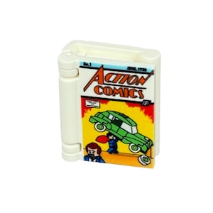 Puede incluir: Un ladrillo Lego blanco con una portada de cómic. La portada muestra el título "Action Comics" con Superman levantando un coche verde. El cómic está sobre un fondo amarillo y rojo.